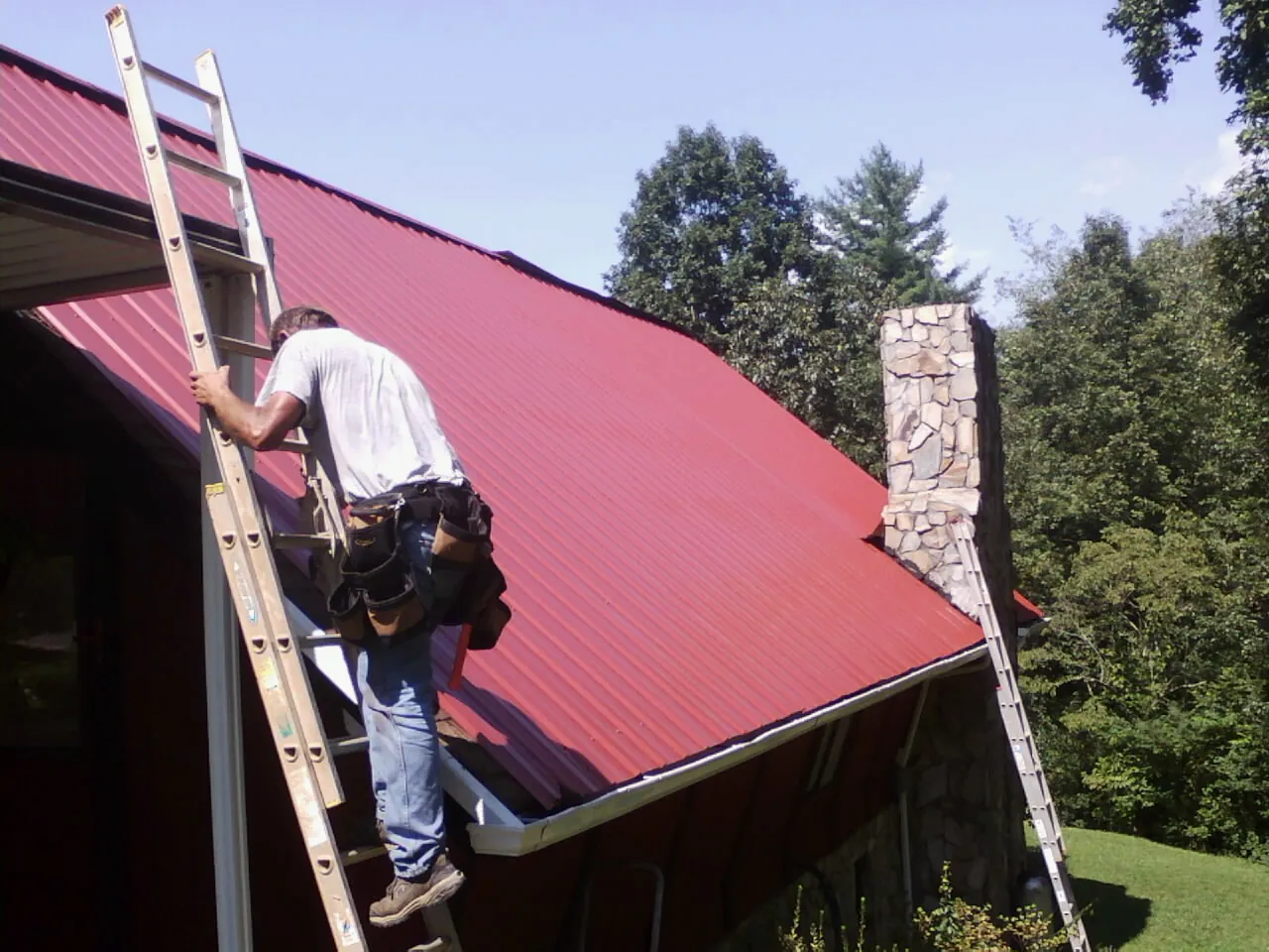 Roofer on a ladder beside a red metal roof and stone chimney — Soffit & Fascia Repair by Roofing Brothers in Louisville
